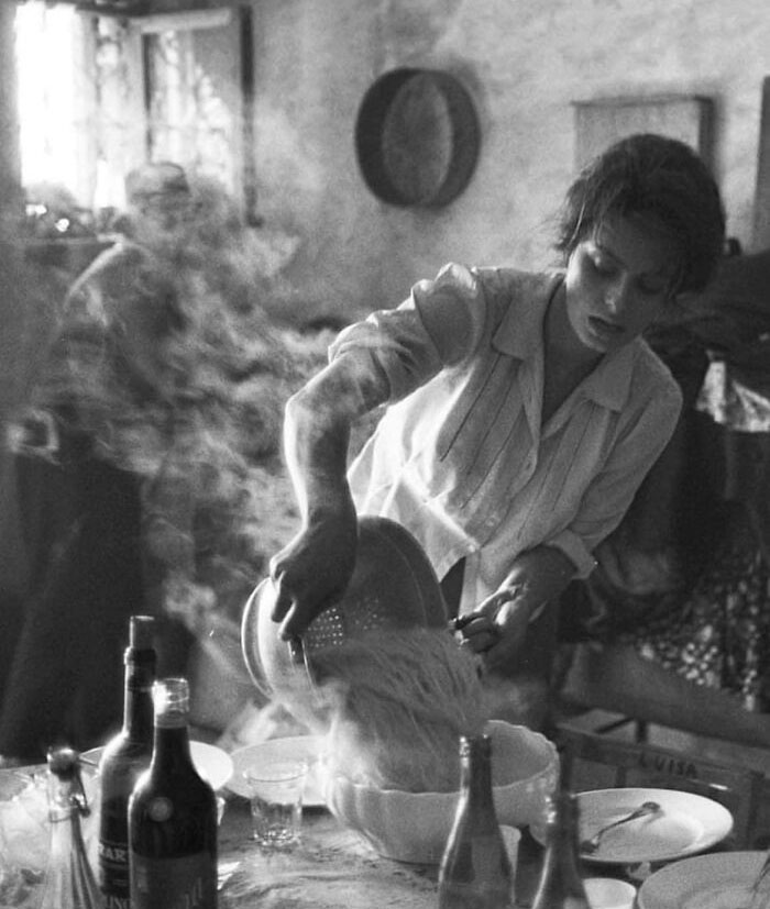 Woman in vintage kitchen draining pasta, steam rising, representing forgotten corners of history.