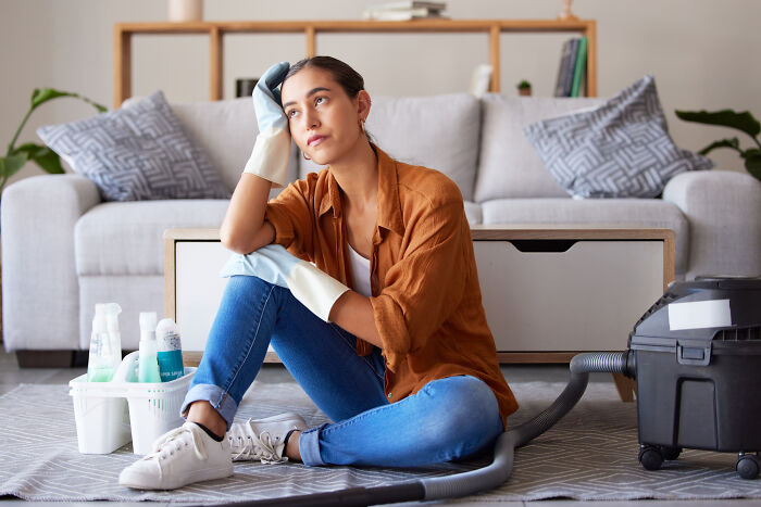 Woman in gloves taking a break from house cleaning, sitting on the floor with vacuum and supplies around her.