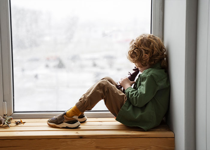 Child sitting alone by a window, suggesting feelings related to "trashy parent" behaviors.