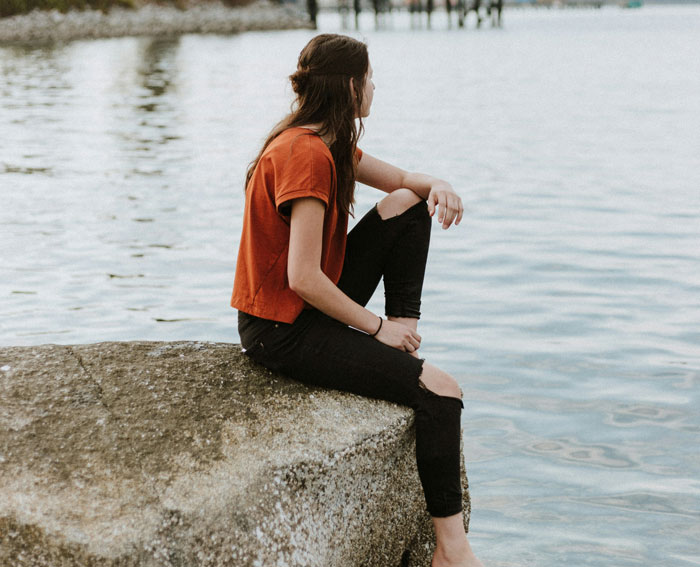 Woman sitting on a rock by the water, reflecting on mental health issues.