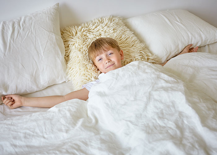 Boy smiling in bed with white sheets and fluffy pillow.
