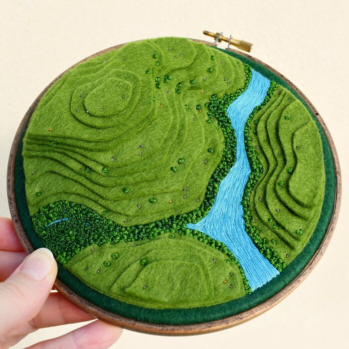 Landscape embroidery featuring green hills and a blue river, showcasing intricate textile art.