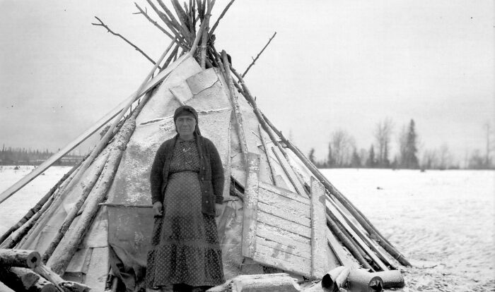 A woman stands in front of a rustic wooden shelter on a snowy landscape, illustrating world changes over the past 100 years.