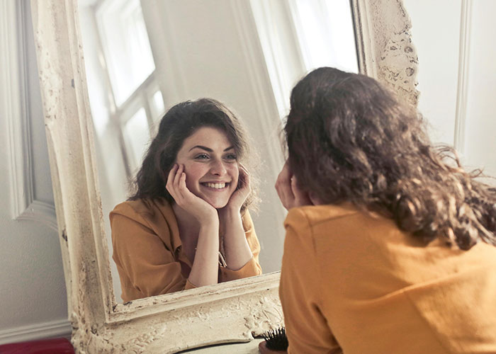 Smiling woman looking in the mirror, practicing beauty tips with a joyful expression.