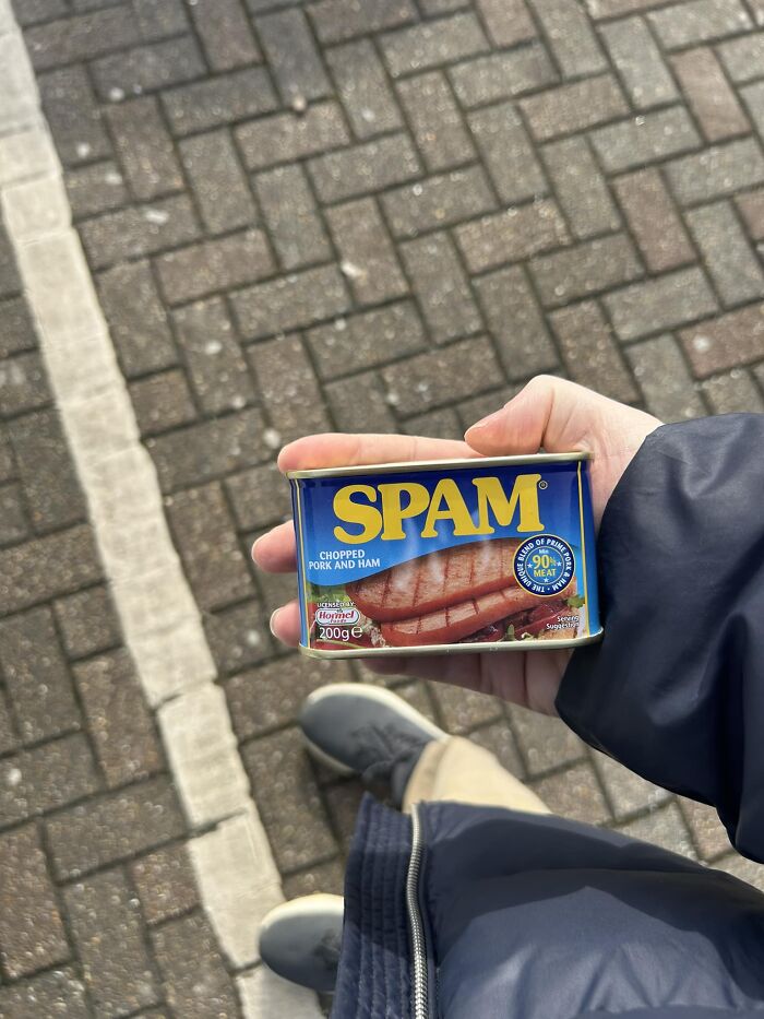 Person holding a can of Spam on a pavement, highlighting British humor.