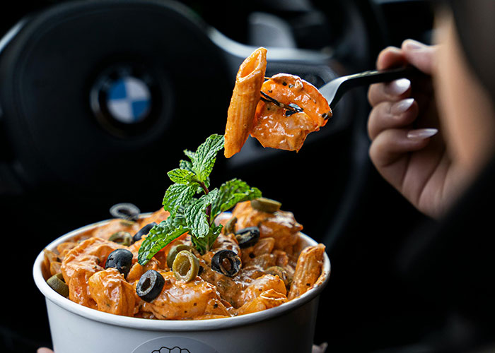 A person eating penne pasta with olives and mint garnish inside a car.