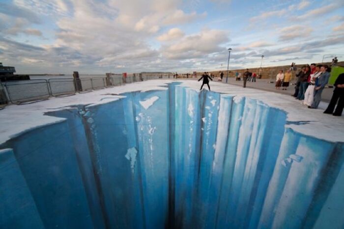 3D street art depicting a deep icy chasm on a sidewalk, with a person playfully balancing at the edge.