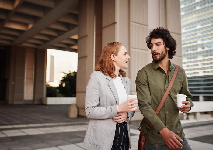 A couple on a first date, walking and talking while holding coffee cups outside a modern building.