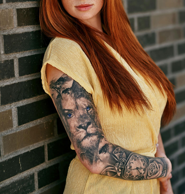 Person with a lion tattoo sleeve on their arm, leaning against a wall, wearing a yellow top.