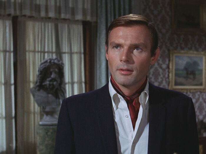 Man in a suit with a red scarf, standing indoors next to a sculpture, illustrating celebrity interactions.