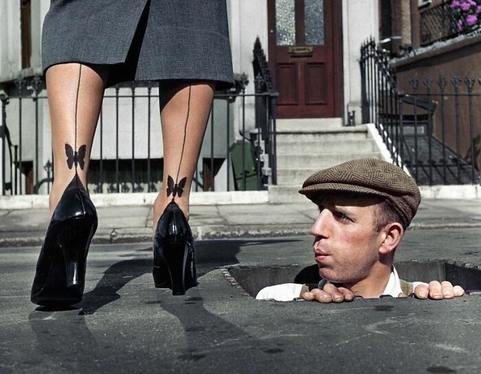 Man peeking from a street hole looks at woman's butterfly-tattooed heels. Perfectly timed street photo capturing humor.