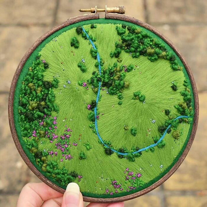 Landscape embroidery art in a hoop, featuring a vibrant green terrain with a winding blue path and scattered floral details.