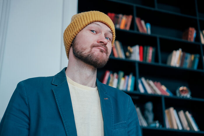 Man in yellow beanie and blue jacket smirking, standing in front of bookshelf.