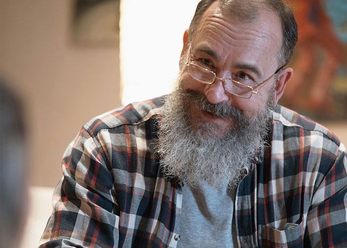 A bearded man in glasses smiles warmly, wearing a plaid shirt, embodying positive reactions to coming out.