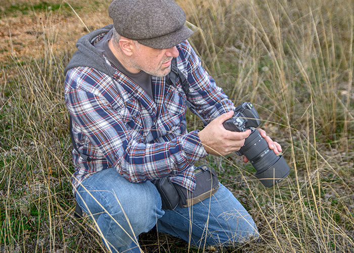 A dad in a plaid shirt and cap crouches in a field, inspecting a camera.