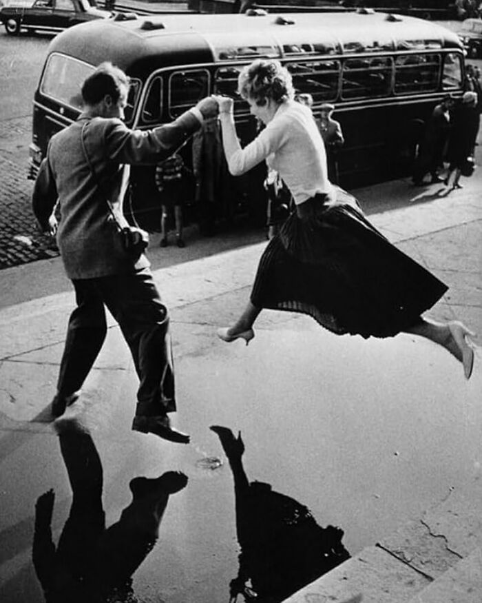 A man and woman leap over a puddle, creating a perfectly timed street photo capturing an unexpected moment.