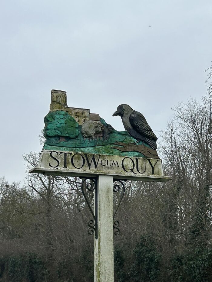 Village sign of Stow c*m Quy featuring a crow and landscape, capturing a humorous British essence.