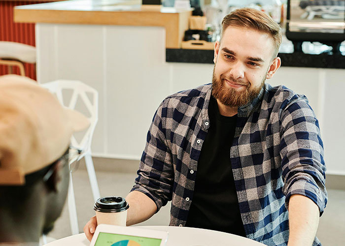 A smiling man in a plaid shirt, sitting at a cafe table, having a supportive conversation with a friend, coffee in hand.