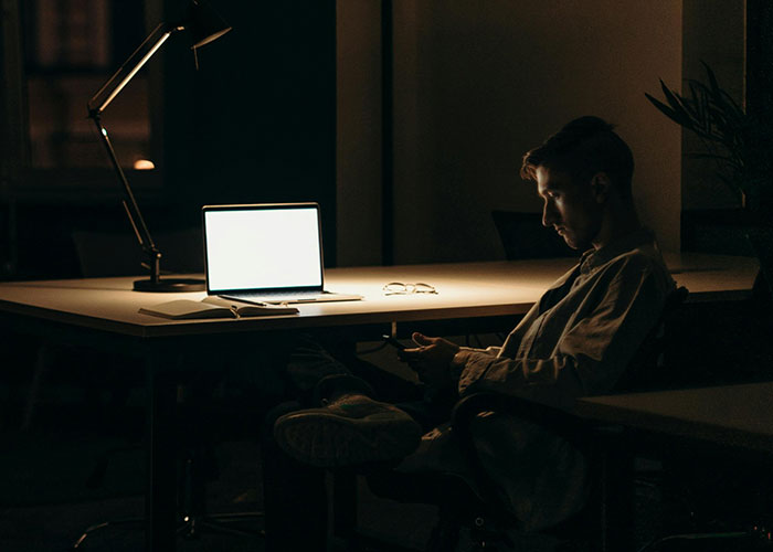 Person sitting in a dimly lit office, focused on a bright laptop, depicting a potentially toxic workplace environment.