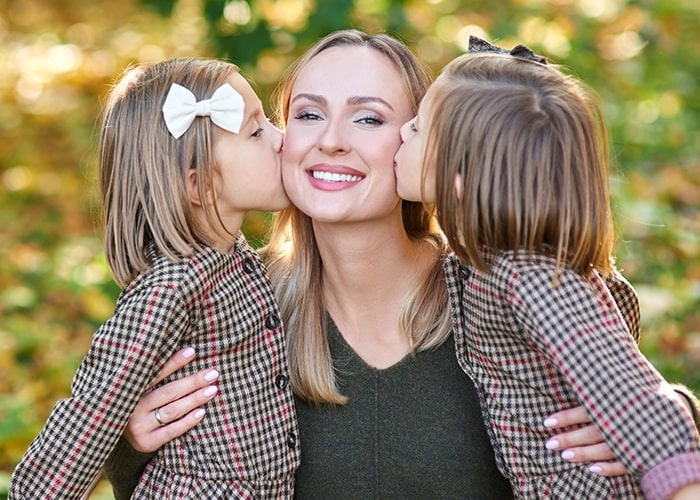 Mother smiling as two daughters kiss her on the cheeks, highlighting parenting moments in a sunny park setting.