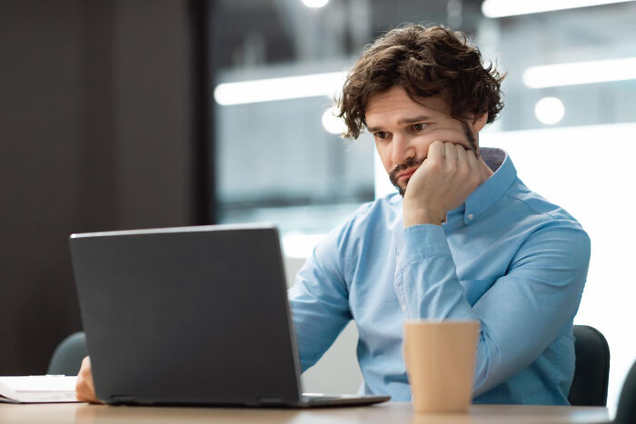 Man in blue shirt looks frustrated at laptop, pondering revenge at work, with coffee cup nearby.