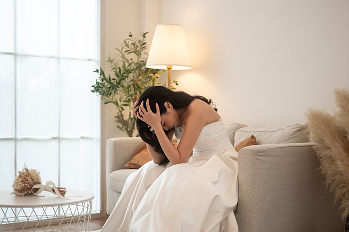 Bride sitting on a sofa, holding her head in distress, wearing a wedding dress in a softly lit room.