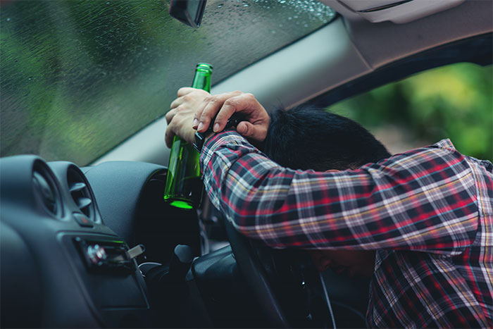 Man in car holding bottle, head on steering wheel, wearing plaid shirt, rainy window visible.