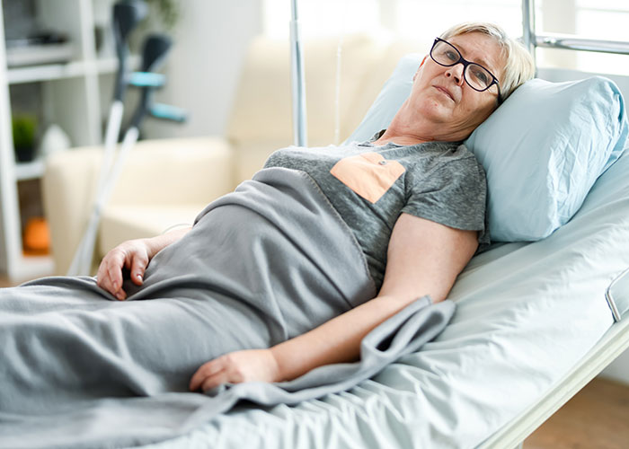 Elderly woman resting in a hospital bed, wearing glasses, covered with a gray blanket.