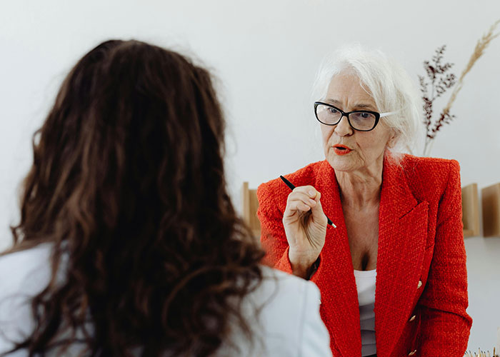 Woman in red blazer pointing a pen, discussing potential workplace toxic traits with a colleague.