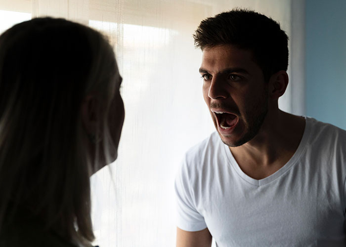 Man yelling at a woman by a window, upset after checking girlfriend's phone, proposal plans ruined.