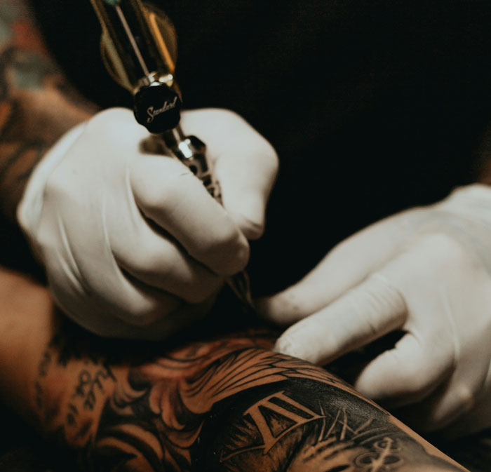 Tattoo artist with gloves creating intricate design on arm, highlighting tattoos' impact on attractiveness.
