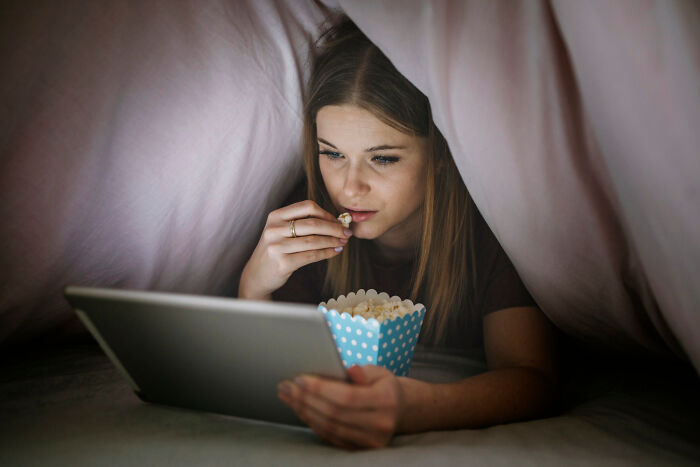 Woman enjoying popcorn under a blanket while watching a tablet, embracing old people habits.