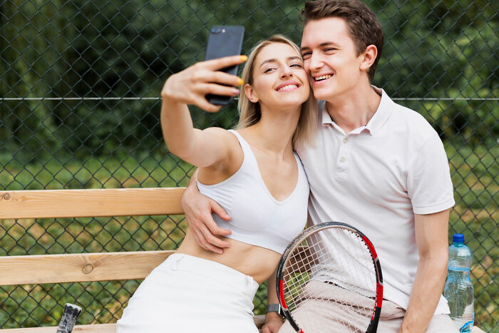 A couple on a first date taking a selfie on a bench, smiling with a tennis racket nearby.