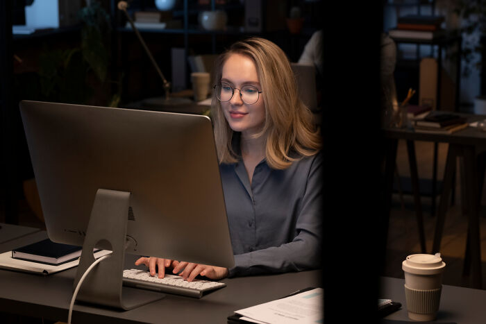 Woman with a "useless" degree working at a computer, wearing glasses and a blue shirt, in an office with a coffee cup nearby.
