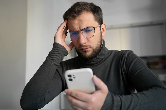 Man in glasses and a dark sweater looks puzzled at his phone, planning harmless April Fools' pranks at home.