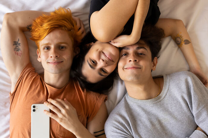 Three friends lying on a bed, smiling at the camera; focuses on relationships.