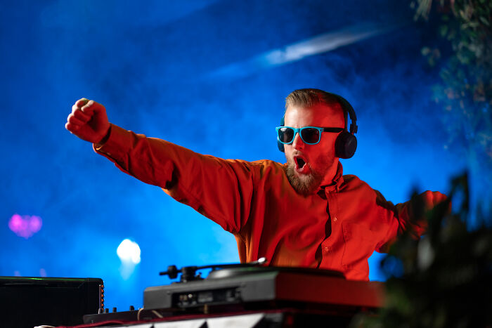 DJ with headphones and sunglasses, energetically playing music on stage, vibrant lights in background.