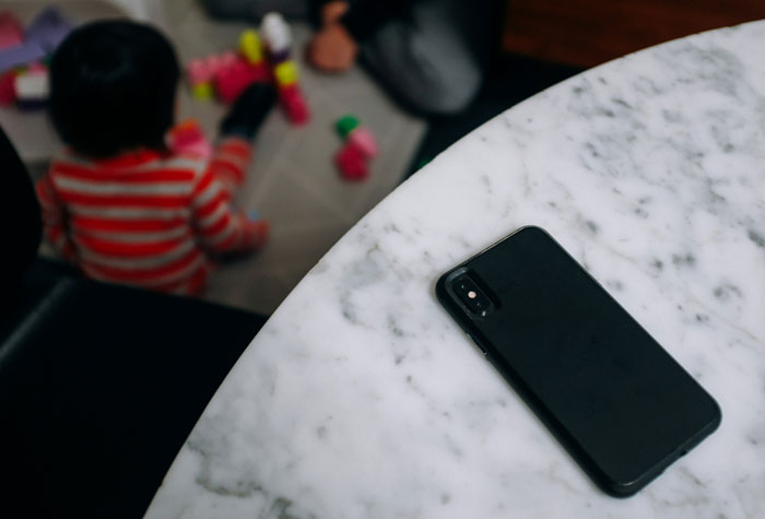 Child playing with blocks on floor, phone on a marble table in focus, reflecting a babysitting scenario for low pay discussion.