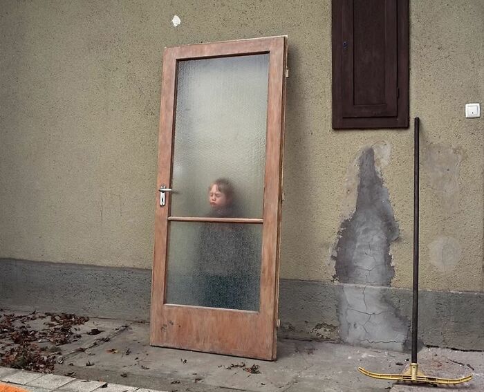 Child peering through a frosted glass door on a weathered street; perfectly timed street photo capturing unexpected moment.