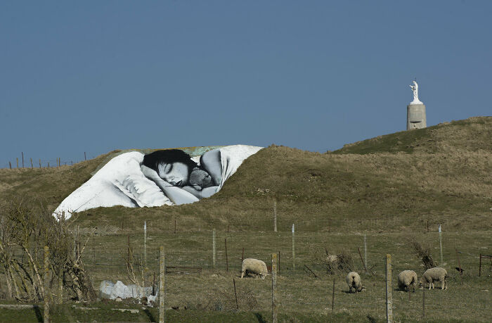 3D street art on a hillside depicting a sleeping figure, surrounded by sheep and a distant statue.