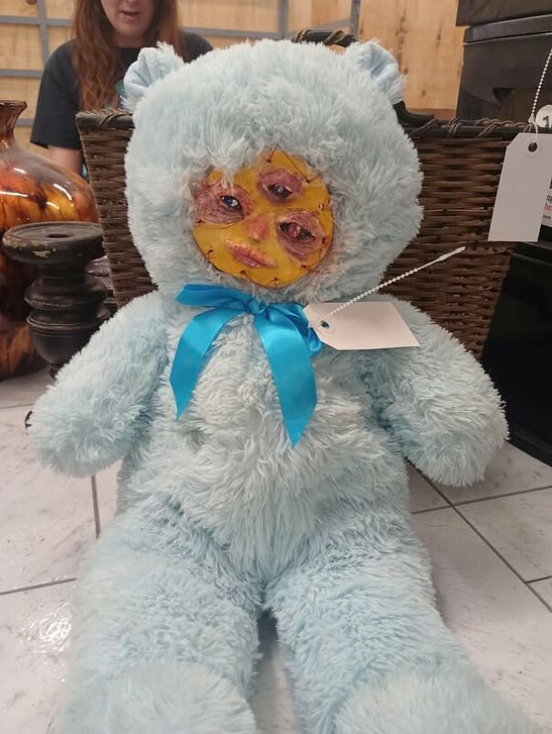 A strange thrifting find: a blue teddy bear with a surreal face, adorned with a blue ribbon and tags, displayed in a store.