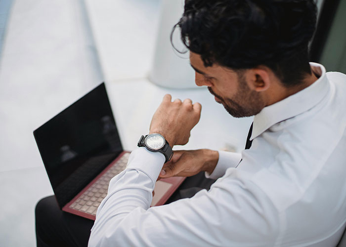 Man in white shirt checking watch, seated with laptop; potential workplace toxic red flag.