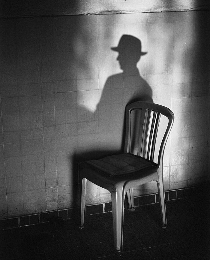 Shadow of a person with a hat beside an empty chair, creating a perfectly timed street photo.