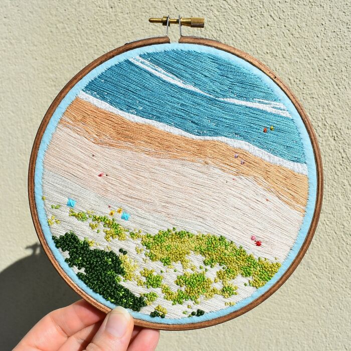 Hand holding a circular embroidery depicting an incredible landscape with ocean, sand, and greenery.