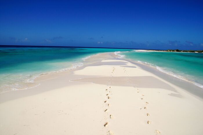 Pristine beach with white sand and turquoise water, footprints leading into the distance under a clear blue sky.