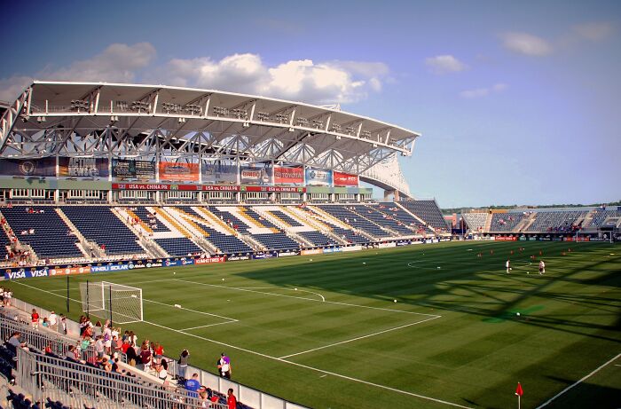 Modern soccer stadium under a blue sky with spectators and players warming up on the green field.
