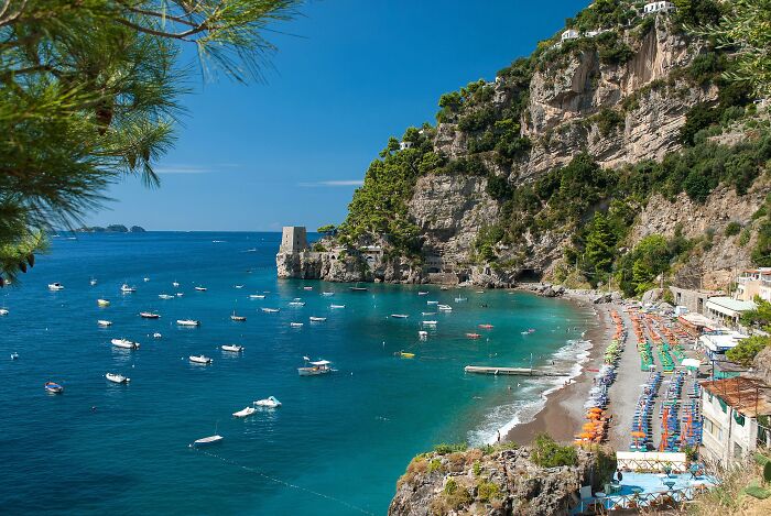 Stunning beach with clear blue waters and boats, surrounded by cliffs and vibrant beach umbrellas.