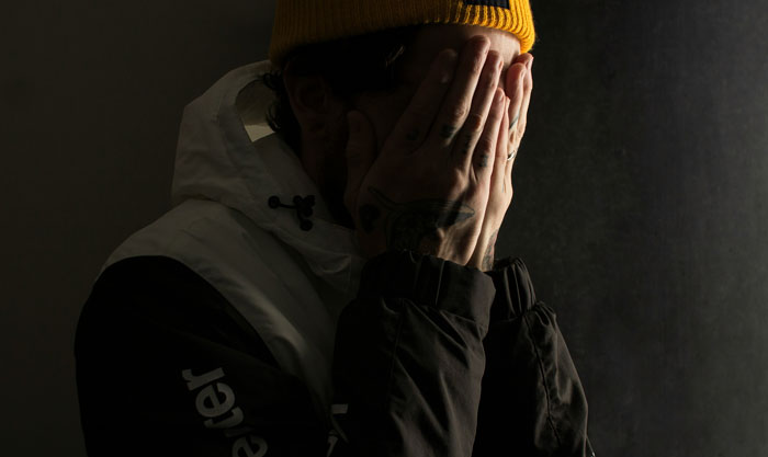 A person with a yellow beanie covers their face with hands, wearing a winter jacket, in a dark, moody setting.