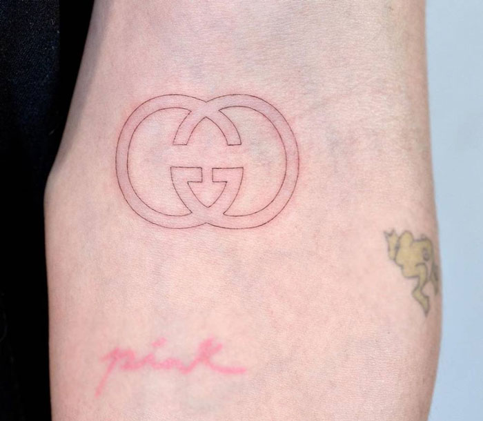 Simple outlined Gucci logo tattoo on skin with two smaller tattoos, including one that says "pink."