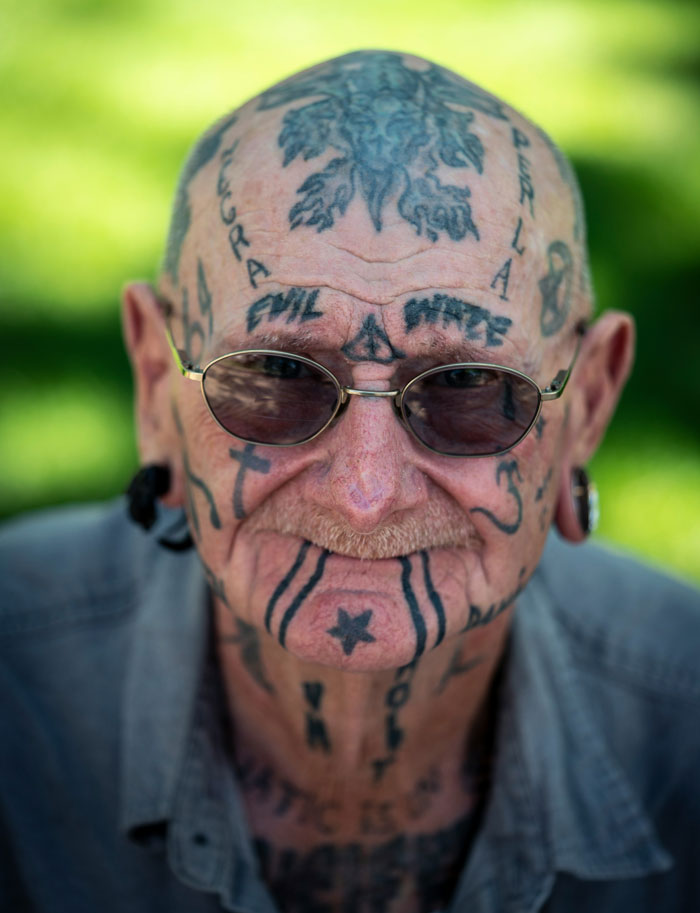 Bald man with facial and head tattoos, wearing sunglasses, showcasing intricate designs.